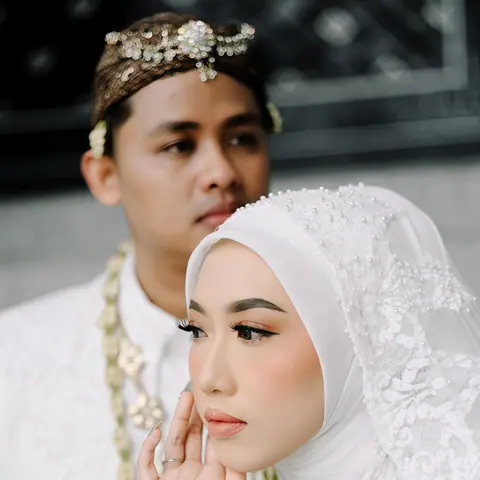 Fadli dan Aini di hari pernikahan: pengantin wanita berhijab putih dengan untaian melati, tangannya di pipi, pengantin pria berbaju putih dengan hiasan kepala bermanik di belakangnya