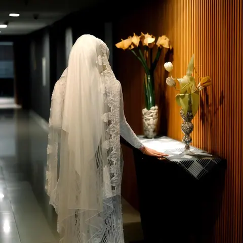 Aini, sang pengantin, dengan veil renda panjang dan gaun, di samping meja bunga di lorong hotel