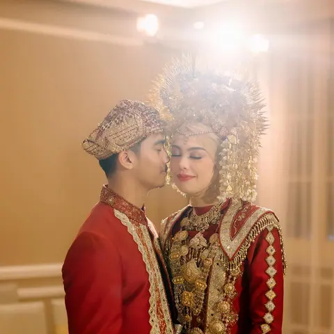 Nadin dan Muiz berbusana merah dan emas, pengantin wanita bermahkota emas tinggi, pengantin pria mencium pelipisnya