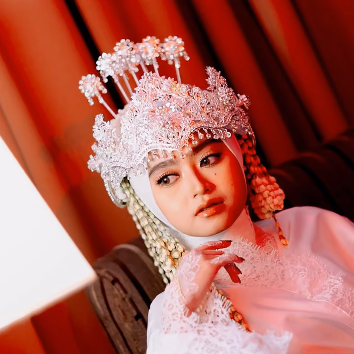 Pengantin wanita di pernikahan Syahla dan Bagas, bermahkota manik merah muda-putih, dagunya bertumpu di tangan, berlatar tirai merah