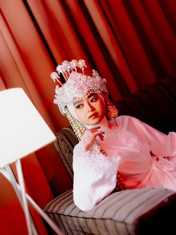 Pengantin wanita bermahkota manik merah muda-putih dan bergaun merah muda, dagunya bertumpu di tangan, berlatar tirai merah